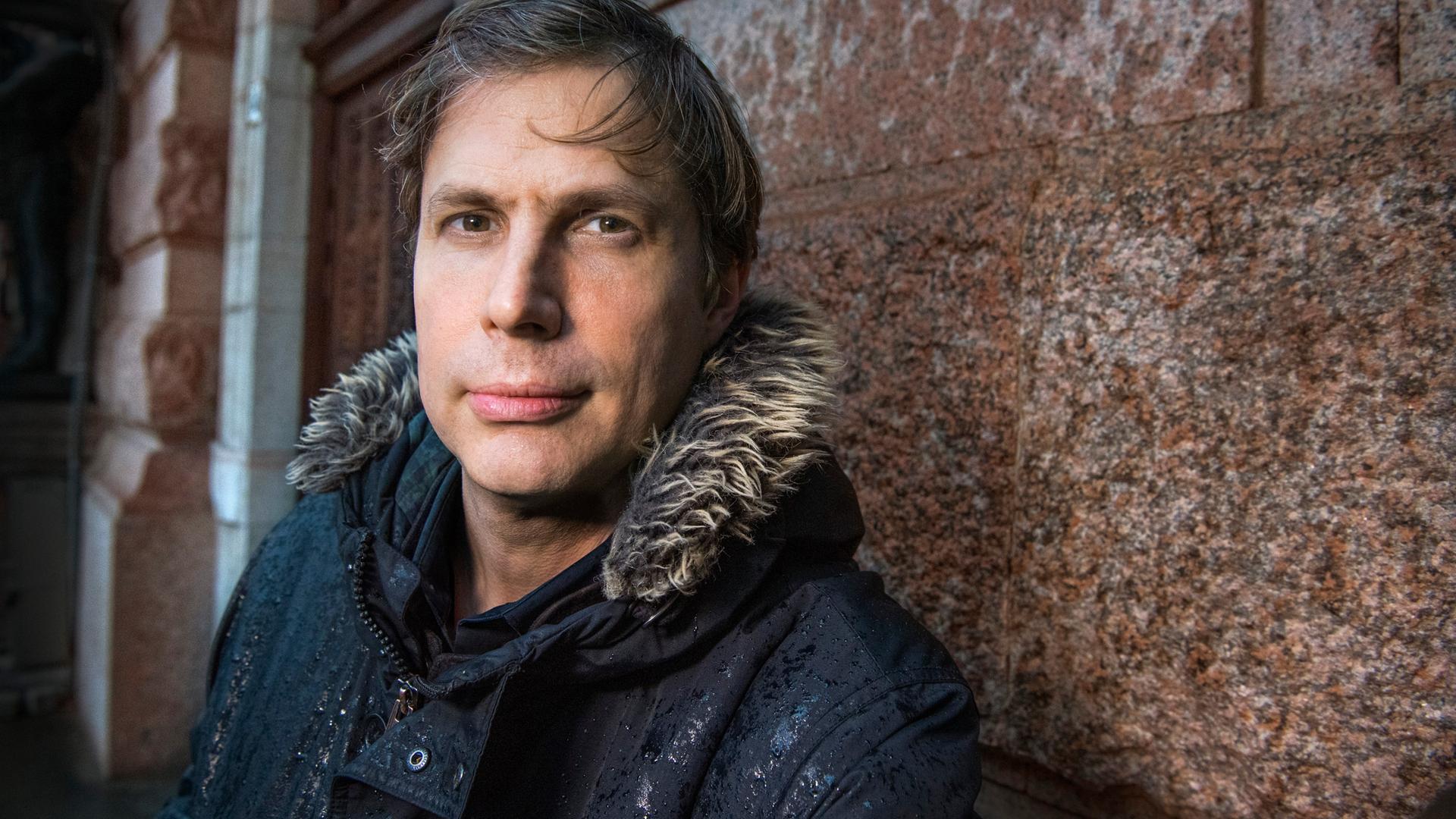 Der Schriftsteller Daniel Kehlmann steht in einer Winterjacke an eine Hauswand gelehnt.