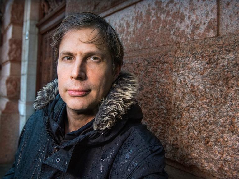 Der Schriftsteller Daniel Kehlmann steht in einer Winterjacke an eine Hauswand gelehnt.