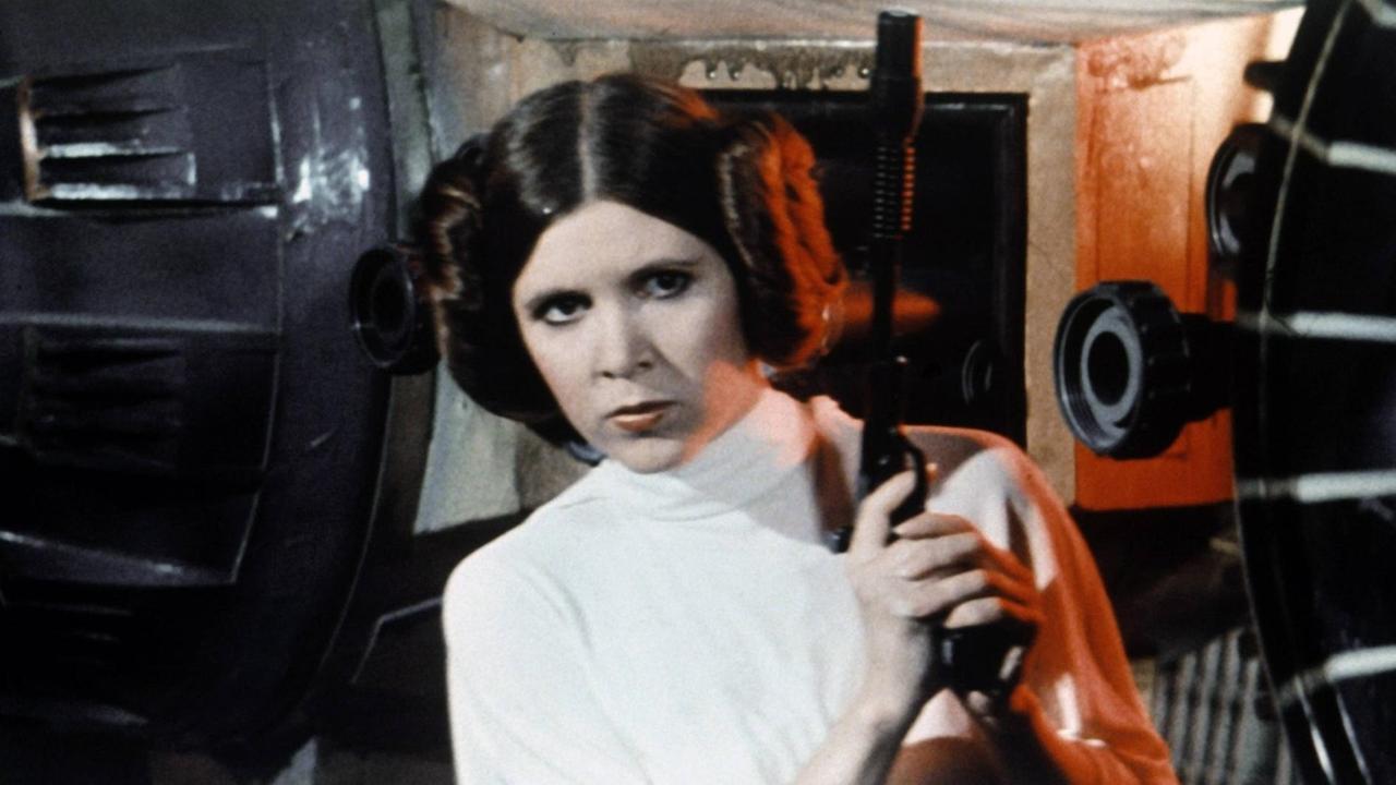 Sie sehen die Schauspielerin Carrie Fisher als "Prinzessin Leia" in "Krieg der Sterne". Sie trägt eine Waffe.