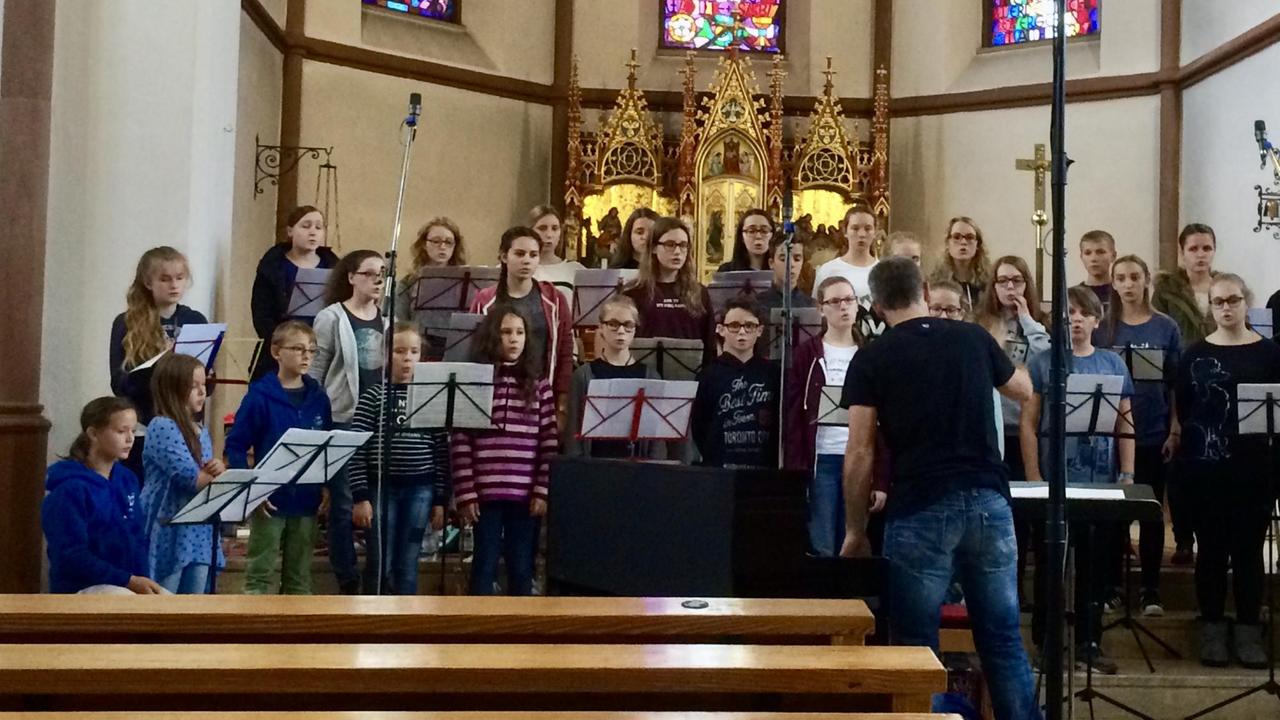 Ein Kinderchor steht in einem Kirchenraum und singt für die um ihn stehenden Mikrofone.