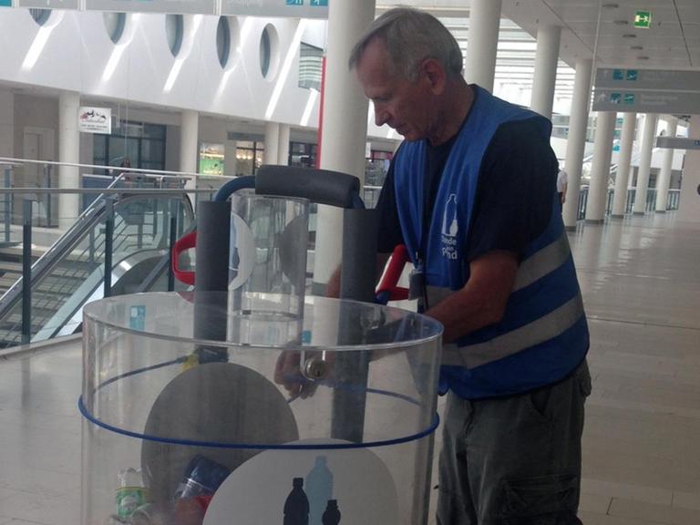 Ein Mann mit blauer Schutzweste zieht einen Plastikbehälter, der gefüllt ist mit leeren Pfandflaschen, durch einen Gang auf einem Flughafen. Spende dein Pfand - heißt die Aktion.