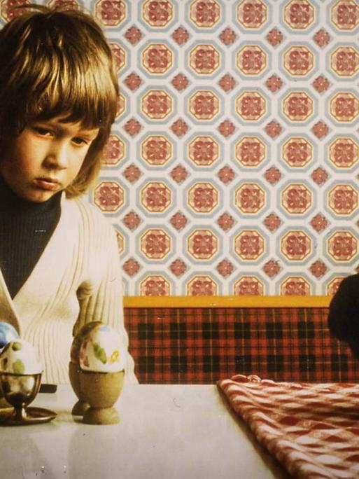 Ein Junge sitzt mit traurigem Gesicht an einem Tisch, auf dem bemalte Ostereier stehen.