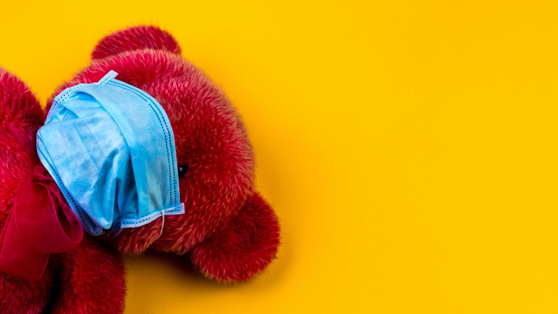 Ein roter Teddybärmit Mundschutz vor gelbem Hintergrund.