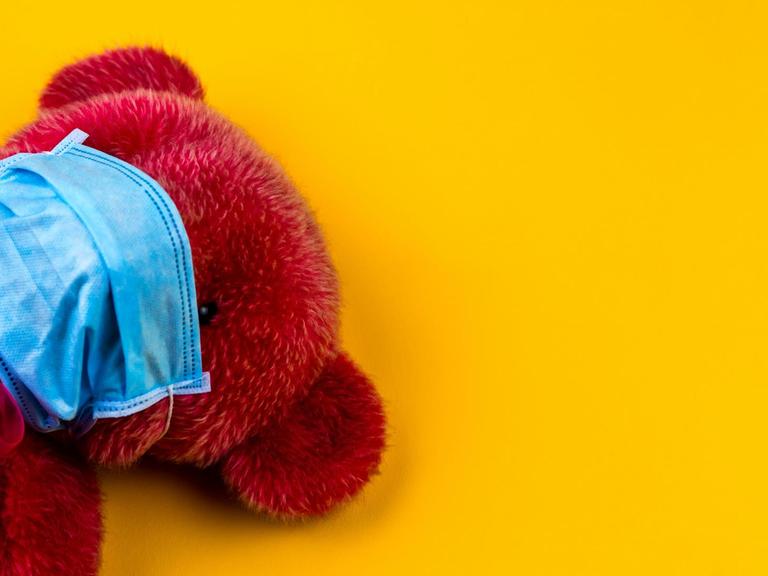 Ein roter Teddybärmit Mundschutz vor gelbem Hintergrund.