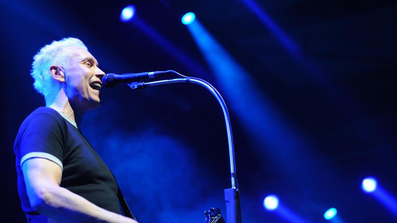 Farin Urlaub ist von der Seite zu sehen, wie er im schwarzen T-Shirt Gitarre spielt und ins Mikrofon singt. Hinter ihm strahlen Scheinwerfer blaues Licht vor schwarzem Hintergrund.