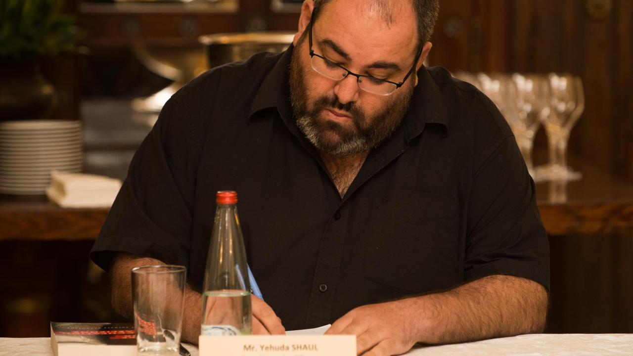 Yehuda Shaul sitzt an einem Konferenztisch und schreibt. Vor ihm ein Namenschild und eine Flasche Wasser.