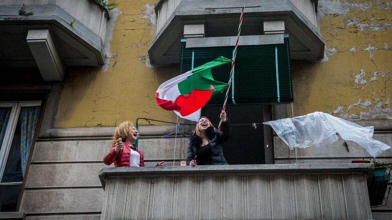 Zwei Frauen stehen singend und lachend auf einem Balkon in Mailand. Eine schwenkt die italienische Nationalflagge.