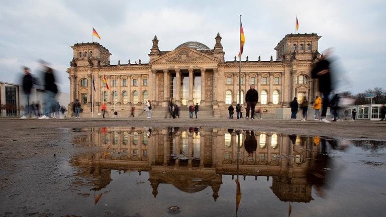 Durch eine Langzeitbelichtung verschwimmen die vorbeigehenden Personen vor dem Bundestag, welcher sich in einer großen Pfütze spiegelt.