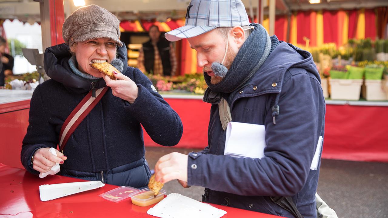 Die Komikerin Cordula Stratmann und der Moderator Olaf Kosert essen Reibekuchen auf dem Klettenbergmarkt. Stratmann erzählt, dass sie hier häufiger herkommt, um die "Rievkooche" für ihren Sohn zu kaufen.