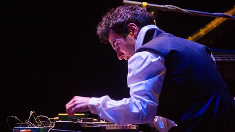 Ein festlich gekleideter Mann spielt in blaues Bühnenlicht gehüllt an einem Keyboard und ist rückseitig zu sehen.