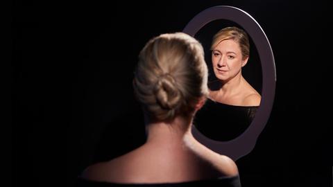 Die Pianistin mit geknoteten Haaren sitzt mit dem Rücken zur Kamera und schaut durch einen Spiegel zurück.