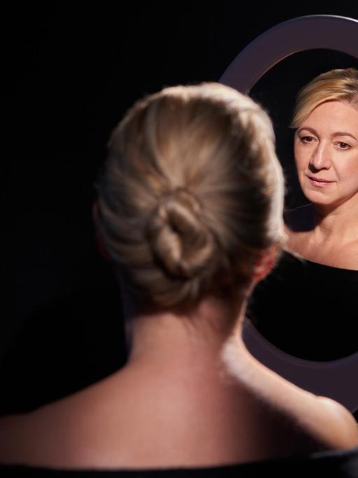 Die Pianistin mit geknoteten Haaren sitzt mit dem Rücken zur Kamera und schaut durch einen Spiegel zurück.