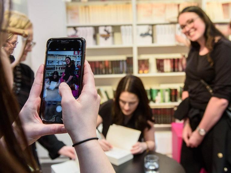 Eine junge Frau fotografiert mit einem Smartphone eine Signierstunde: Autorin Mona Kasten signiert, ein weiblicher Fan lächelt in die Kamera.