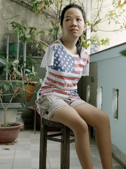 Eine junge Frau sitzt auf einem Hocker auf einem Balkon. Sie hat keine Arme und trägt ein T-Shirt mit aufgedruckter US-Flagge.