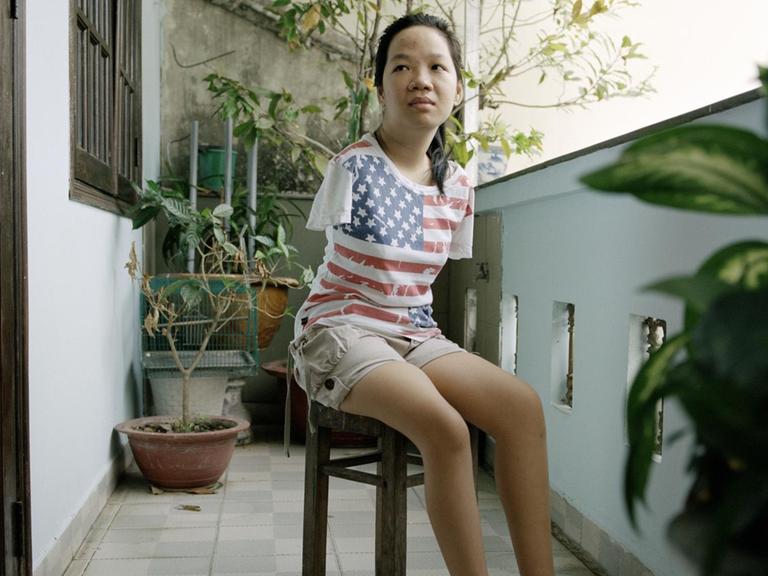 Eine junge Frau sitzt auf einem Hocker auf einem Balkon. Sie hat keine Arme und trägt ein T-Shirt mit aufgedruckter US-Flagge.