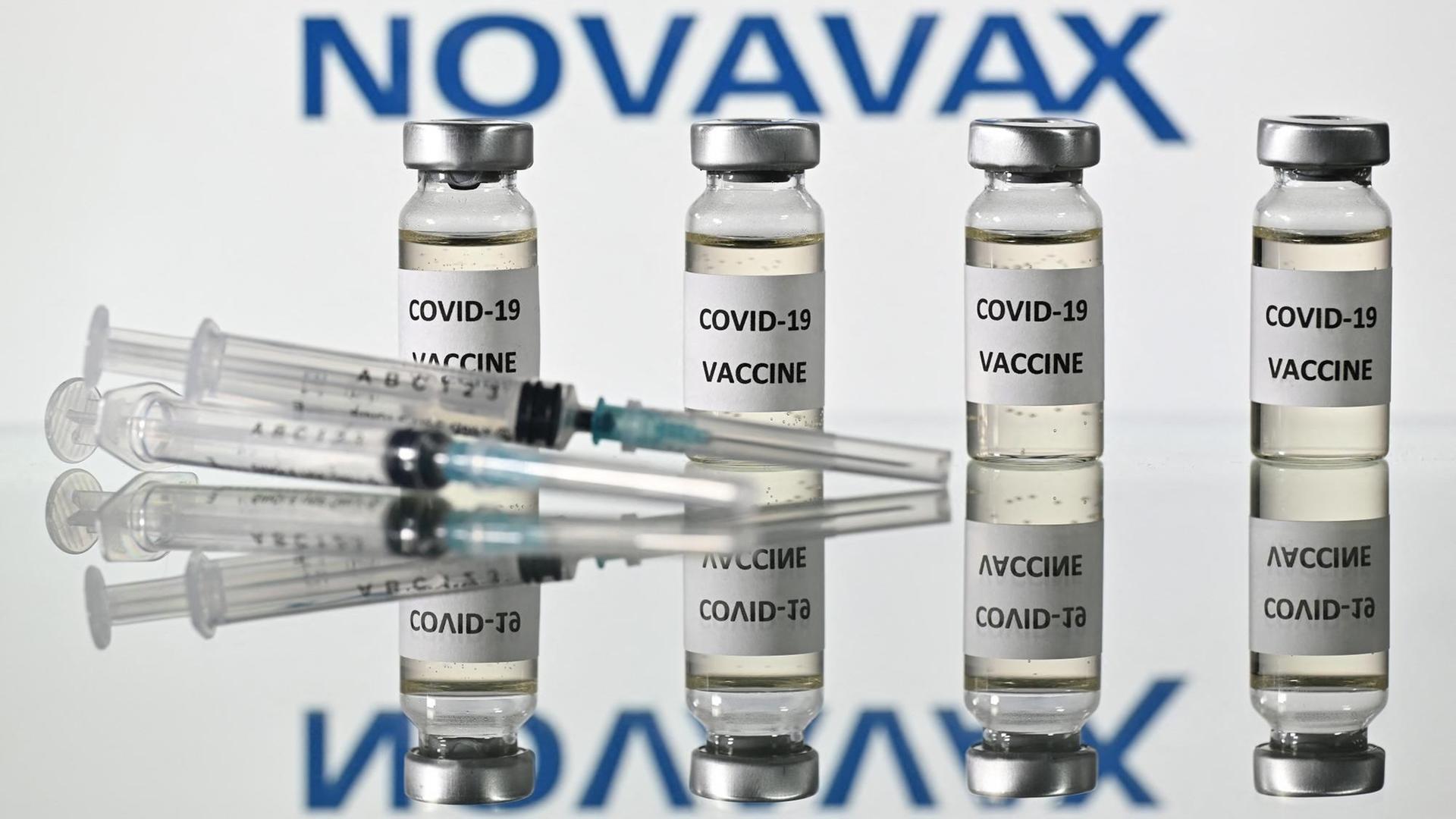 Zwei Spritzen liegen vor vier Ampullen gefüllt mit dem Impfstoff des Herstellers Novavax. Sein Firmenlogo prangt im Hintergrund.