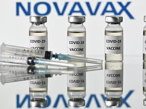 Zwei Spritzen liegen vor vier Ampullen gefüllt mit dem Impfstoff des Herstellers Novavax. Sein Firmenlogo prangt im Hintergrund.
