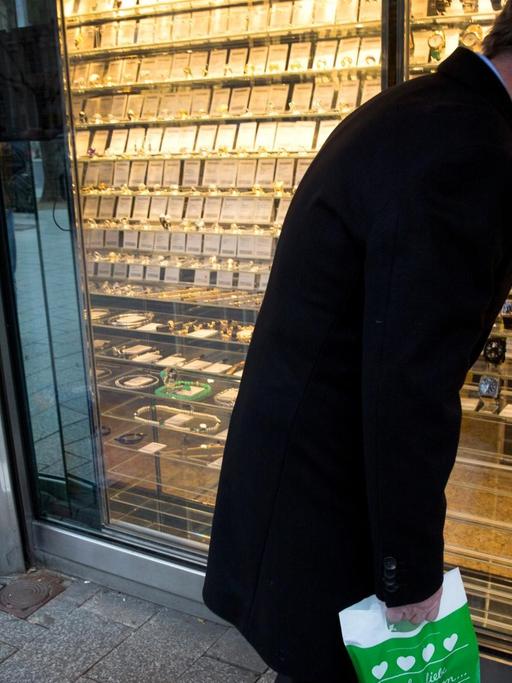 Ein Mann schaut in Düsseldorf auf der Königsallee in ein Schaufenster mit Luxusuhren, während neben ihm ein Bettler um Geld bittet.