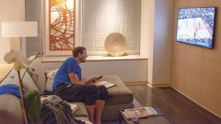 Dirk Nowitzki sitzt auf der Couch in einem Hotelzimmer und schaut ein NBA-Spiel auf einem Fernseher.