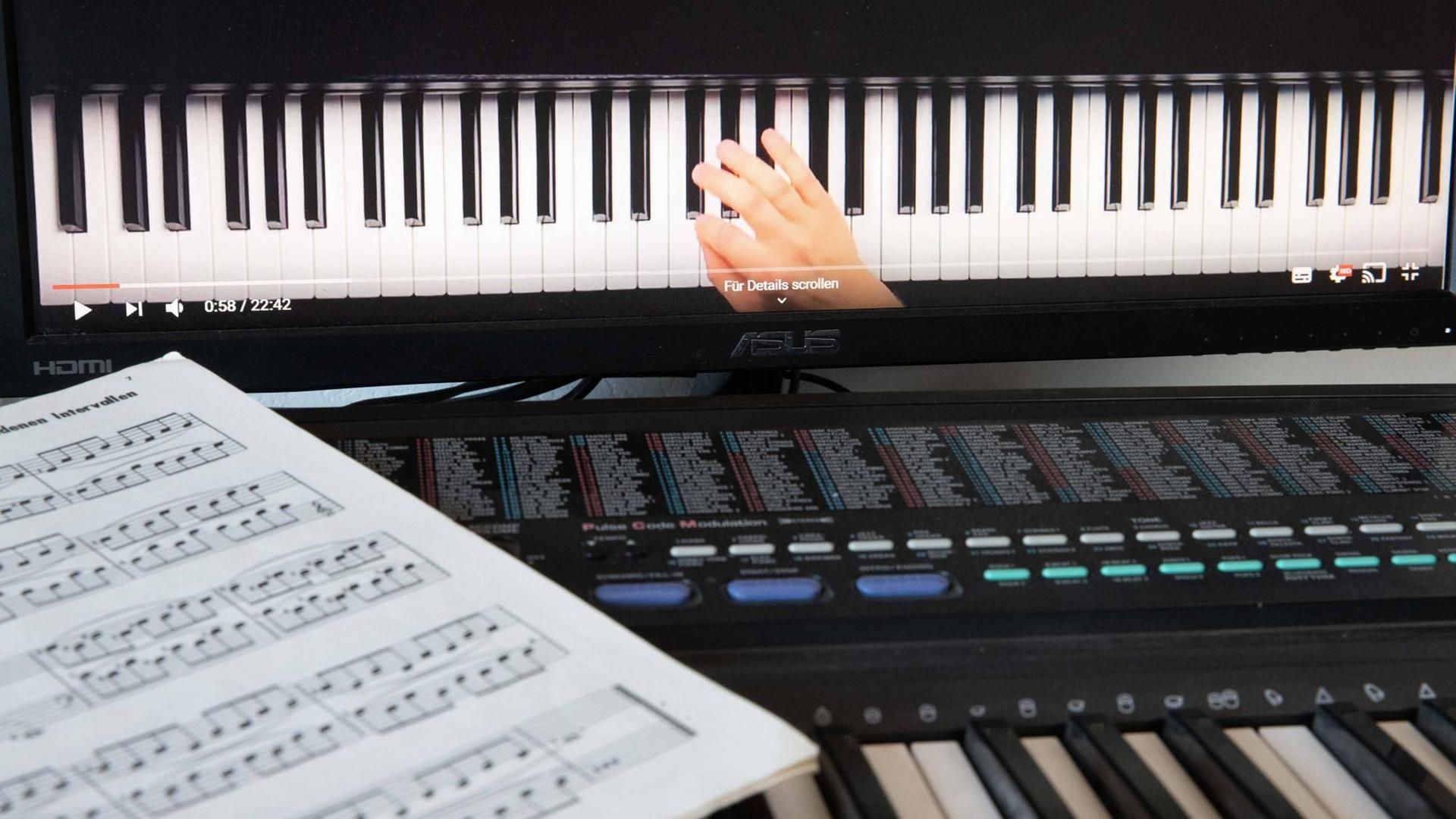 Musikschulunterricht via Skype: Ein Notenblatt liegt auf einem Keyboard, dahinter ist ein Bildschirm aufgebaut, auf dem eine Hand auf einer Klaviertastatur zu sehen ist.