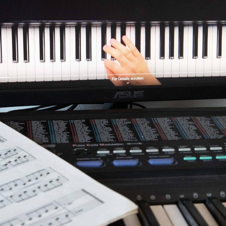 Musikschulunterricht via Skype: Ein Notenblatt liegt auf einem Keyboard, dahinter ist ein Bildschirm aufgebaut, auf dem eine Hand auf einer Klaviertastatur zu sehen ist.