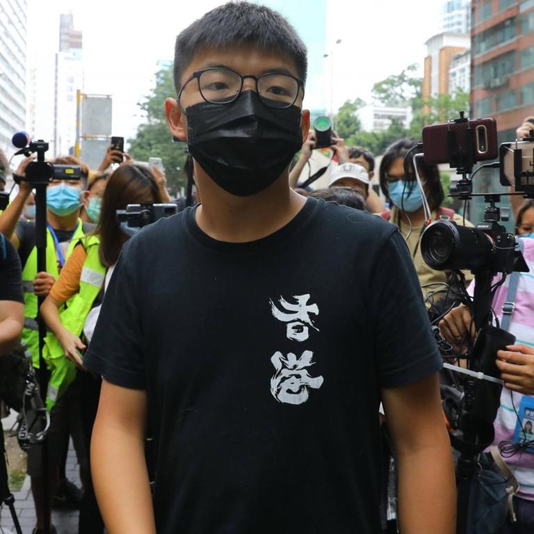 Ein junger Mann mit Brille, schwarzem T-Shirt und schwarzer Mund-Nasen-Maske steht umringt von Journalist*innen.