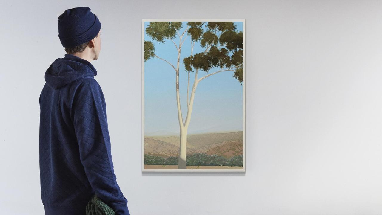 Ein Mann blickt auf ein Bild eines Baumes, das an einer leeren, weißen Wand hängt. Man sieht ihn von hintern. Er trägt eine dunkelblaue Mütze und einen dunkelblauen Pullover.