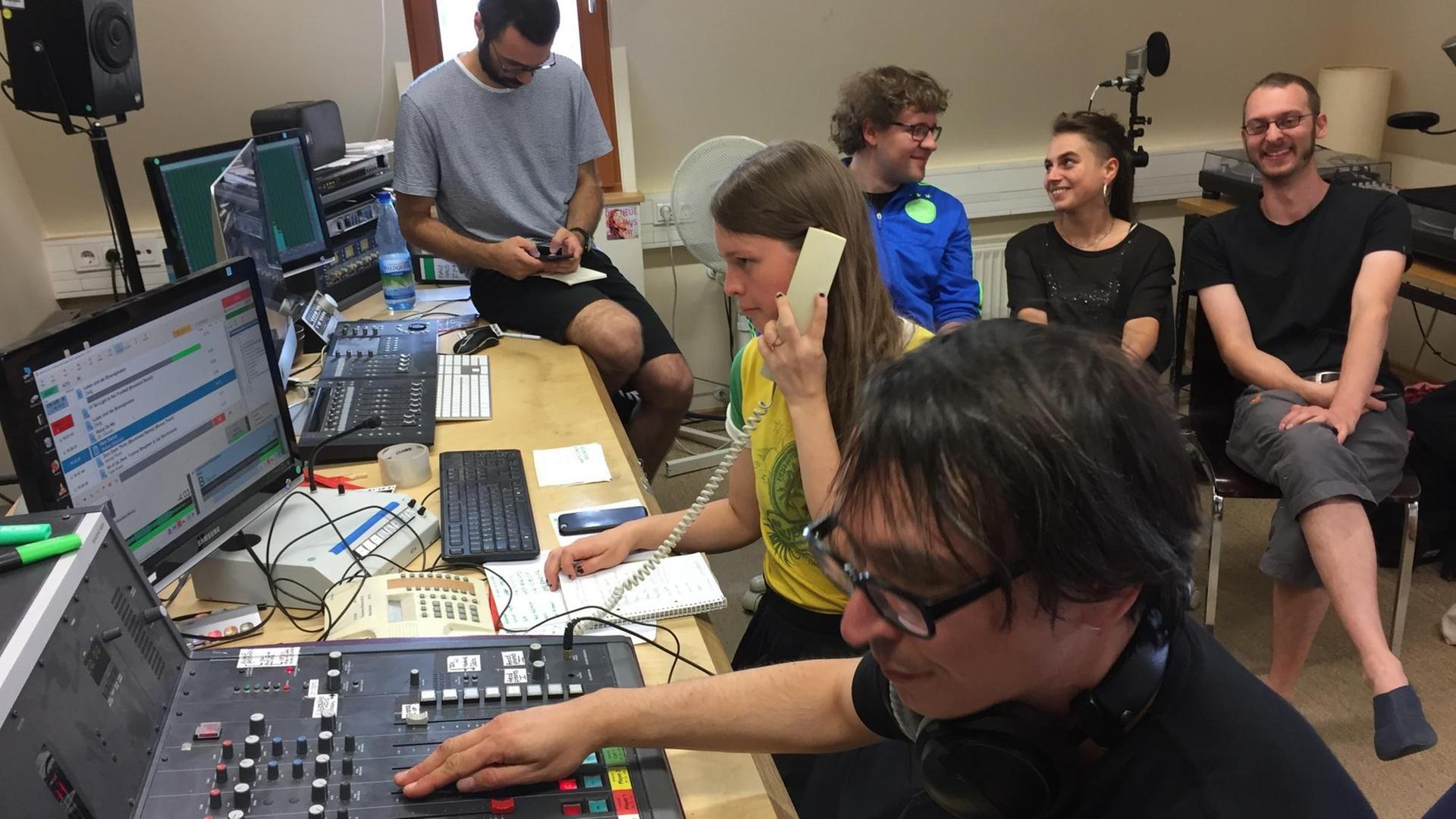 Sechs junge Menschen sitzen in einem Radiostudio, ein Student bedient das Mischpult während eine Studentin telefoniert