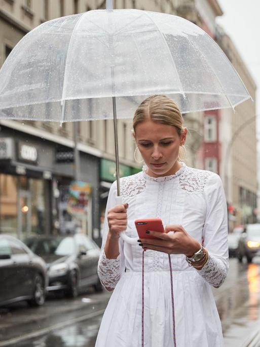 Nena Schink mit einem durchsichtigen Regenschirm und in weißem Kleid in Berlin. Sie tippt auf dem Smartphone. Im Hintergrund ist der Fernsehturm zu erkennen.