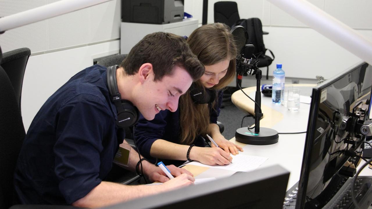 Ein junger Mann und eine junge Frau sitzen im Radiostudio und notieren etwas auf Papier.