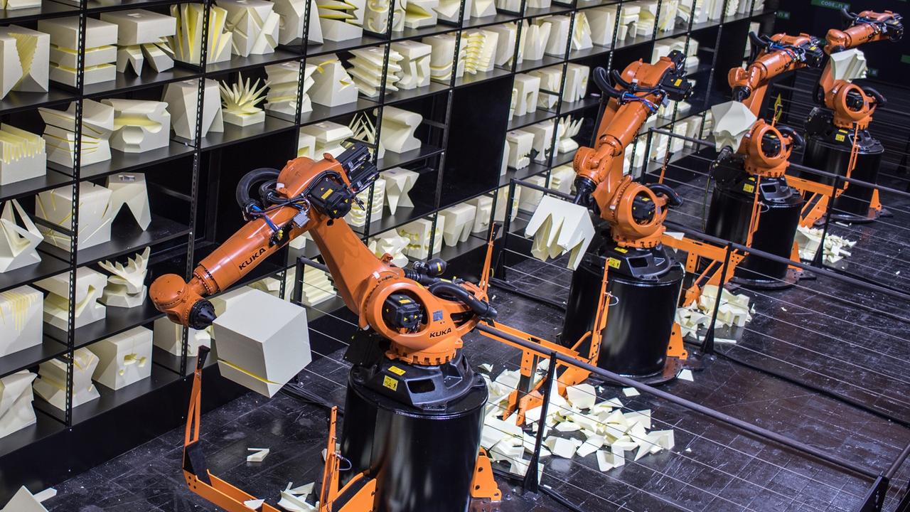 Orangefarbener Produktionsroboterarm, im Hintergrund ein Regal mit Kunstobjekten aus Holz.