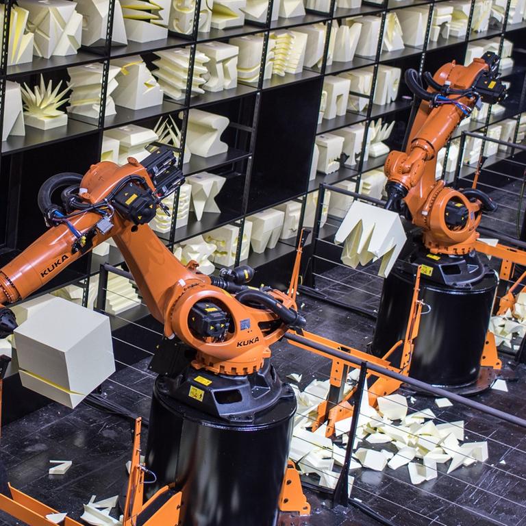 Orangefarbener Produktionsroboterarm, im Hintergrund ein Regal mit Kunstobjekten aus Holz.