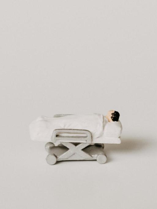 Kleine Modellfigur eines Patienten, der vor weißem Hintergrund in einem Krankenbett liegt.