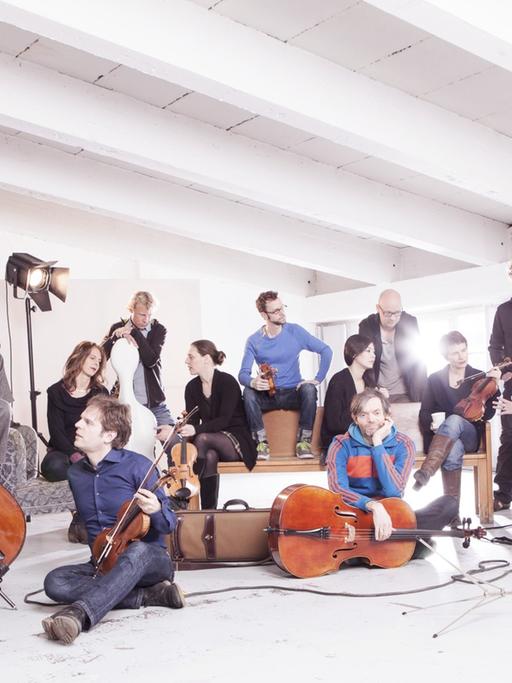Das Ensemble sitzt und steht mit seinen Instrumenten in einem weißen Raum mit markanter Deckenkonstrukion.