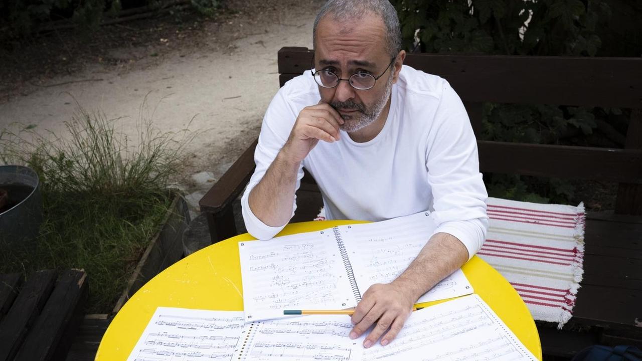 Ein Mann mit Brille sitzt an einem gelben, runden Gartentisch voller Notenblätter.