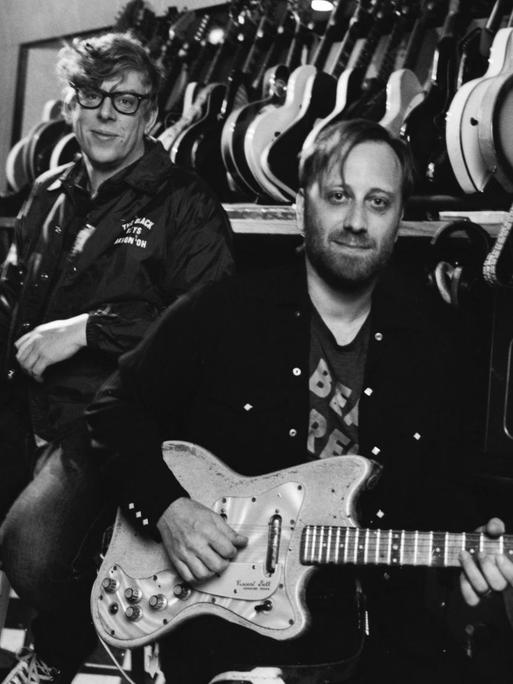 Die beiden Musiker stehen vor einer Wand an der einige Gitarren hängen und schauen in die Kamera.