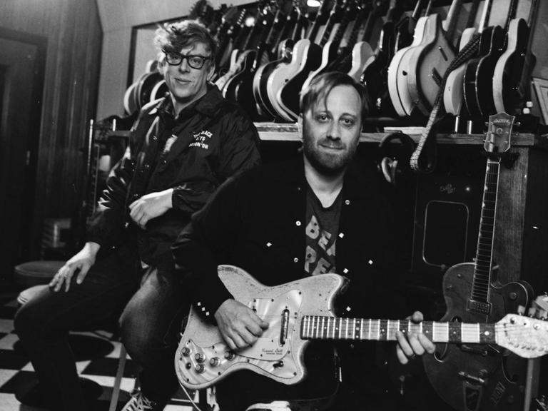Die beiden Musiker stehen vor einer Wand an der einige Gitarren hängen und schauen in die Kamera.
