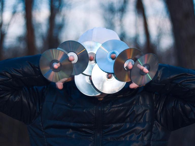 Eine Person hält sich acht CDs vor das Gesicht. Sie steht mitten im Wald.