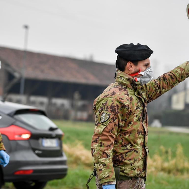 Ein Soldat mit Atemschutzmaske hält an einer Straßensperre vor der roten Zone von Turano Lodigiano eine Winkerkelle in die Höhe. 