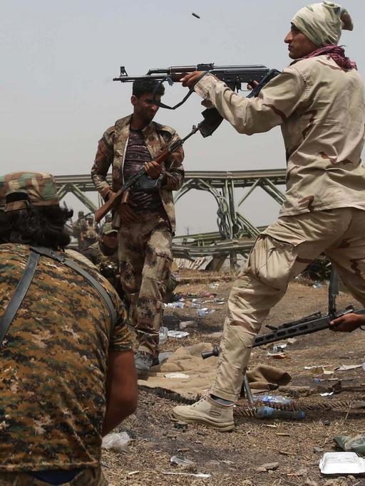 Kämpfer einer irakischen Einheit an einem Flussufer: Einer schießt über eine Böschung auf ein nicht sichtbares Ziel, zwei Andere kauern in Deckung. Im Hintergrund nähern sich weitere Soldaten.