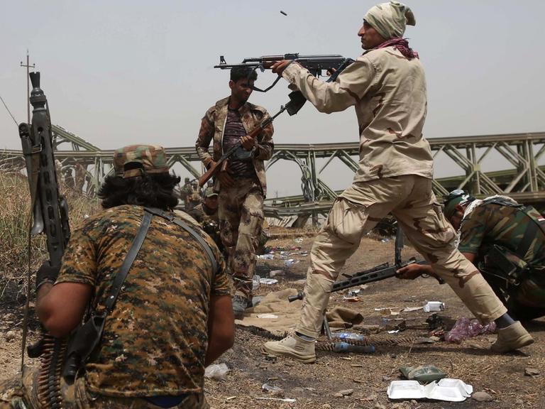 Kämpfer einer irakischen Einheit an einem Flussufer: Einer schießt über eine Böschung auf ein nicht sichtbares Ziel, zwei Andere kauern in Deckung. Im Hintergrund nähern sich weitere Soldaten.