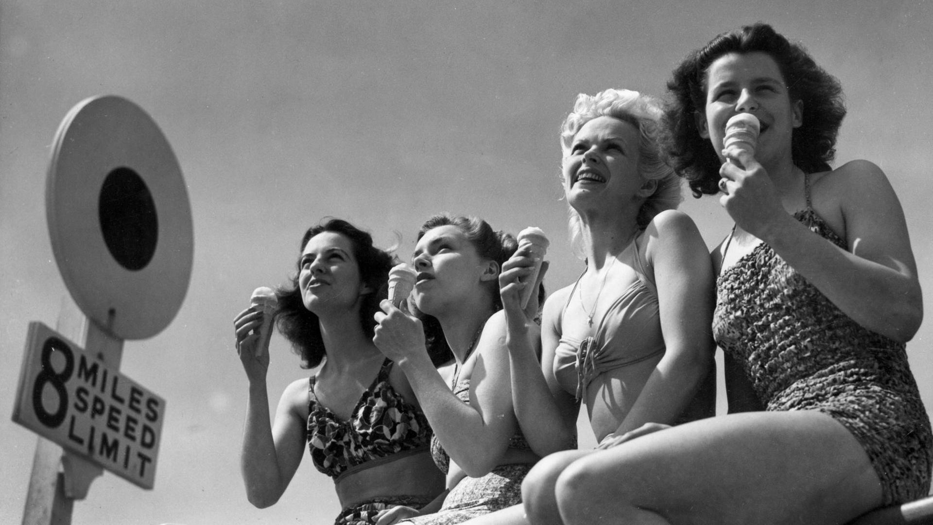 Eine historische Schwarz-Weiß-Aufnahme von 1947 zeigt vier junge Frauen in zeitgenössischer Bikini-Bademode, die an der Strandpromenade von Bournemouth in England ein Eis essen.