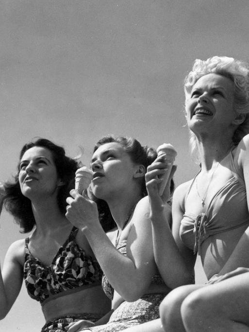 Eine historische Schwarz-Weiß-Aufnahme von 1947 zeigt vier junge Frauen in zeitgenössischer Bikini-Bademode, die an der Strandpromenade von Bournemouth in England ein Eis essen.