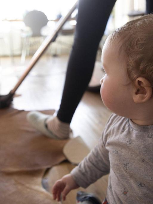 Eine Frau saugt Staub mit einem Staubsauger in ihrem Wohnzimmer. Im Vordergrund sitzt ein Baby auf dem Boden.
