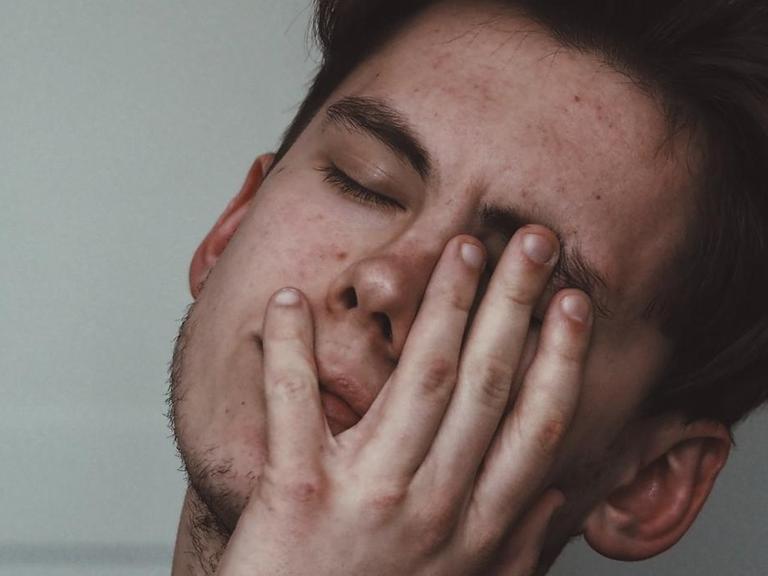 Ein erschöpfter junger Mann legt sein Gesicht mit geschlossenen Augen in seine Hand