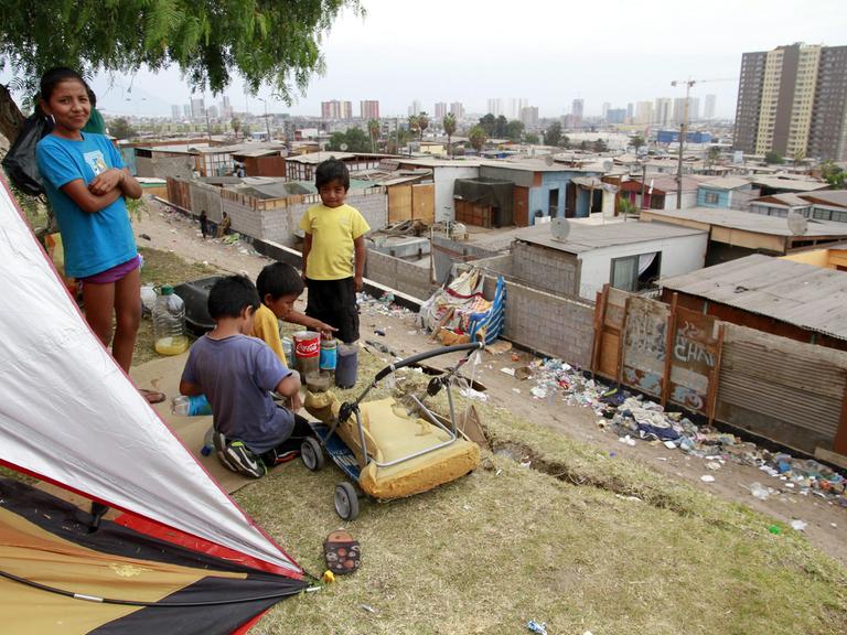 Kinder sitzen im Vordergrund vor einem Zelt und blicken über Ziegel- und Wellblechhütten auf Hochhäuser im Hintergrund.