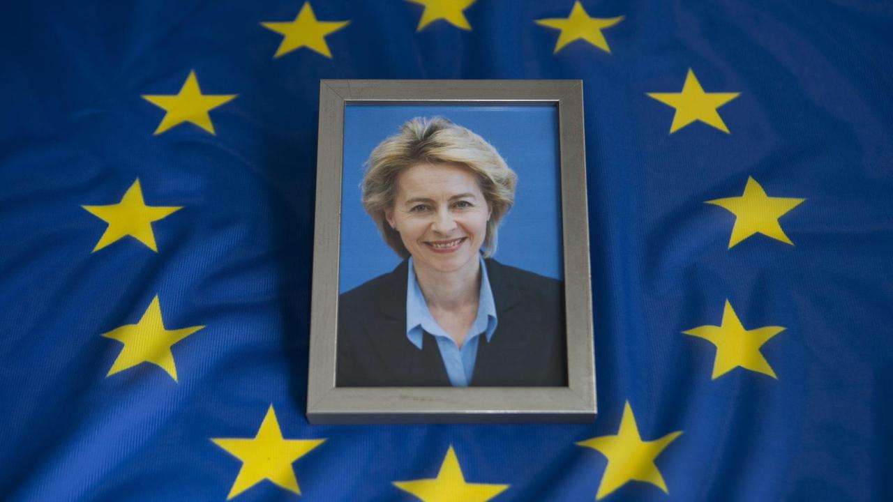 Das Bild zeigt ein gerahmtes Foto von der Politikerin Ursula von der Leyen. Es liegt auf einer blauen Europa-Flagge.