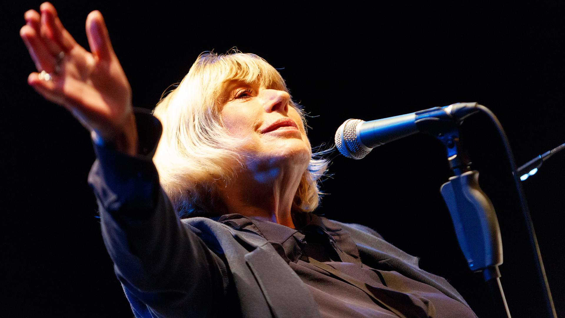 Die englische Sängerin und Songwriterin Marianne Faithfull steht am Mikrofon und winkt mit einer Hand ins Publikum.