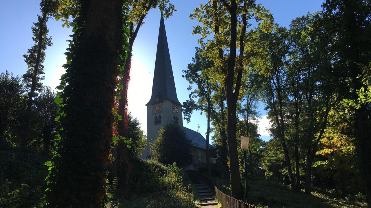 Vor strahlend blauem Himmel erhebt sich eine Kirche mit spitzem Turmdach - im Vordergrund führt ein Weg zwischen Bäumen den Hang hinauf zur Kirche hin.