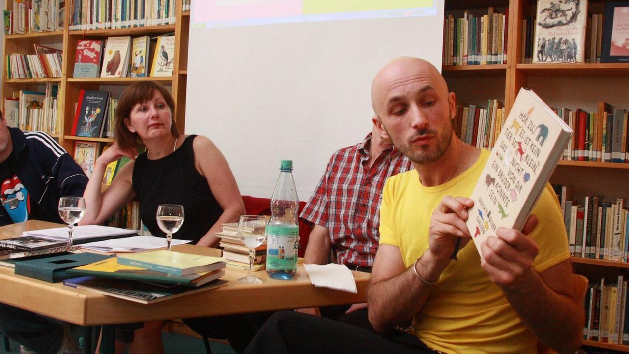 Farbfoto eines jungen Mannes mit Glatze, der ein Buch zeigt bei einer Lesung, es ist der Illustrator Aljoscha Blau